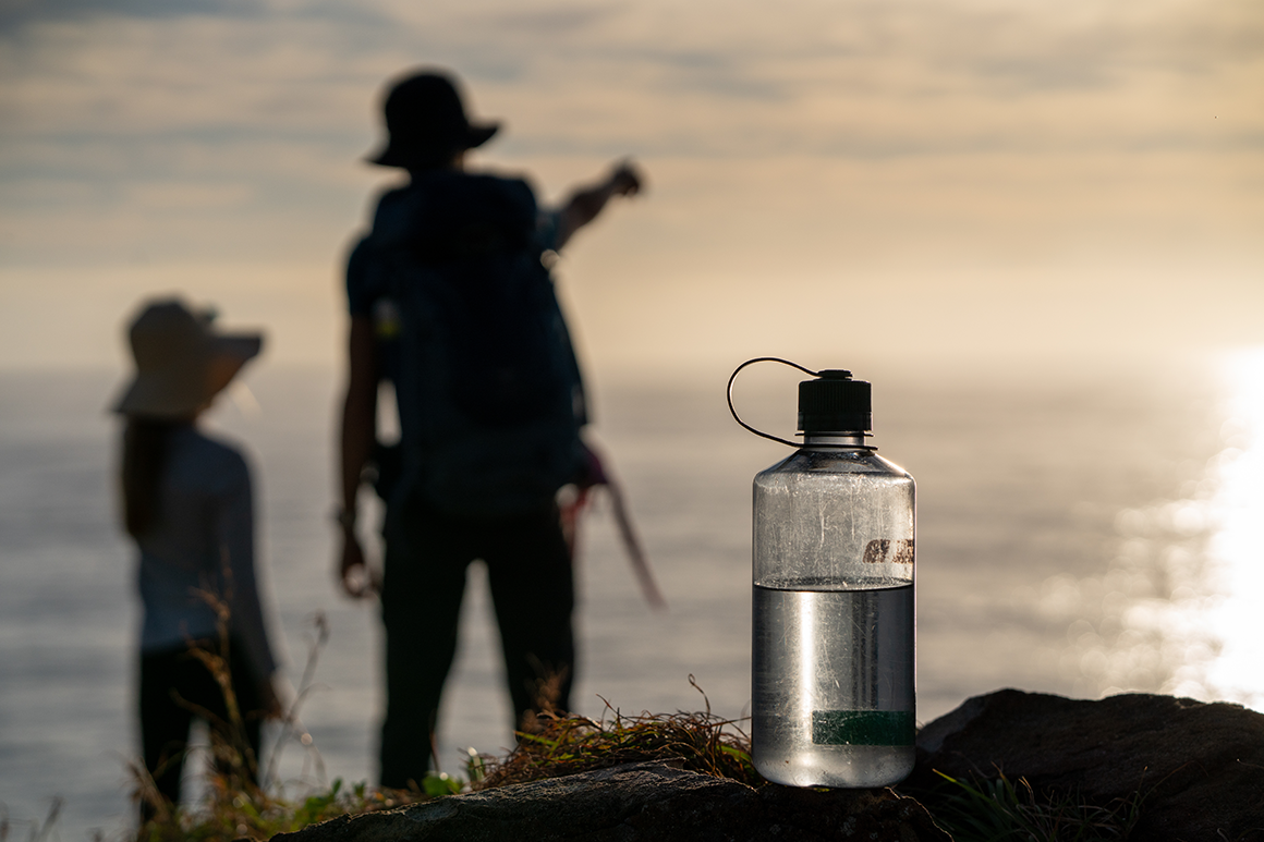 Man and child hikers silhouetted against sunny skies with drink bottle in foreground. 
