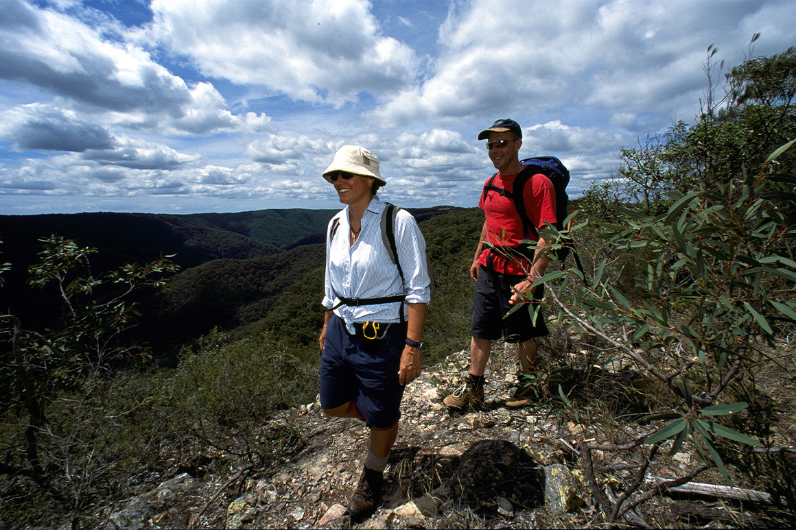 Two walkers traverse a ridge track against backdrop of distant mountains.