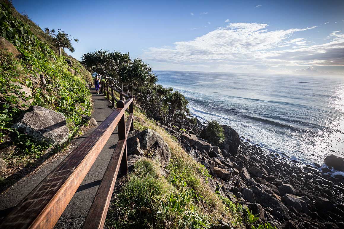 On a steep cliff that drops to a rocky shore and the ocean’s edge, a person walks along sealed track (with handrail) that winds around the cliff-face.    