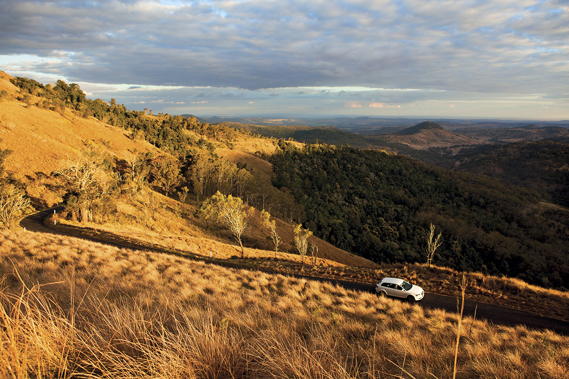 Distant mountain range lies in shadow under a cloudy blue sky and a white car travels along a road winding around a grassy hillside, bathed in the golden glow of late afternoon light.