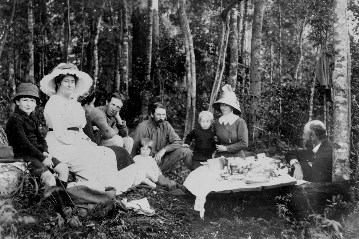 Old black and white image of family of 8 sitting on the ground in a forest around a picnic spread, wearing clothing of the period.