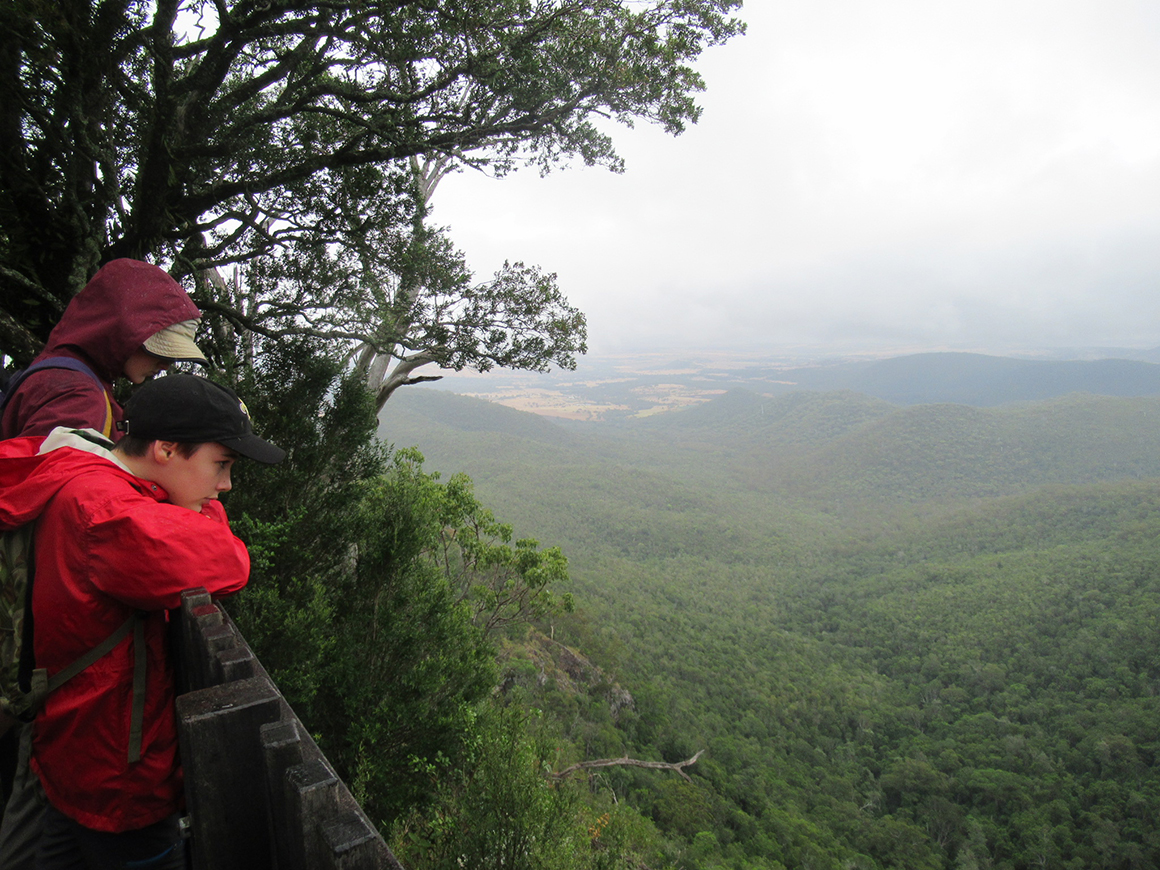 Two young teens, clad in bright red jackets, take a break from hiking, gazing over a handrail at expansive views of forest-clad ranges and valleys under cloudy skies. 