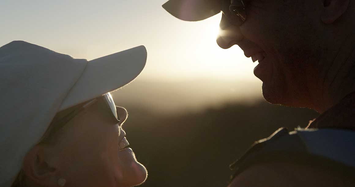 Close up of a couple’s faces (man and woman) smiling at each other, silhouetted against a pale golden sunrise