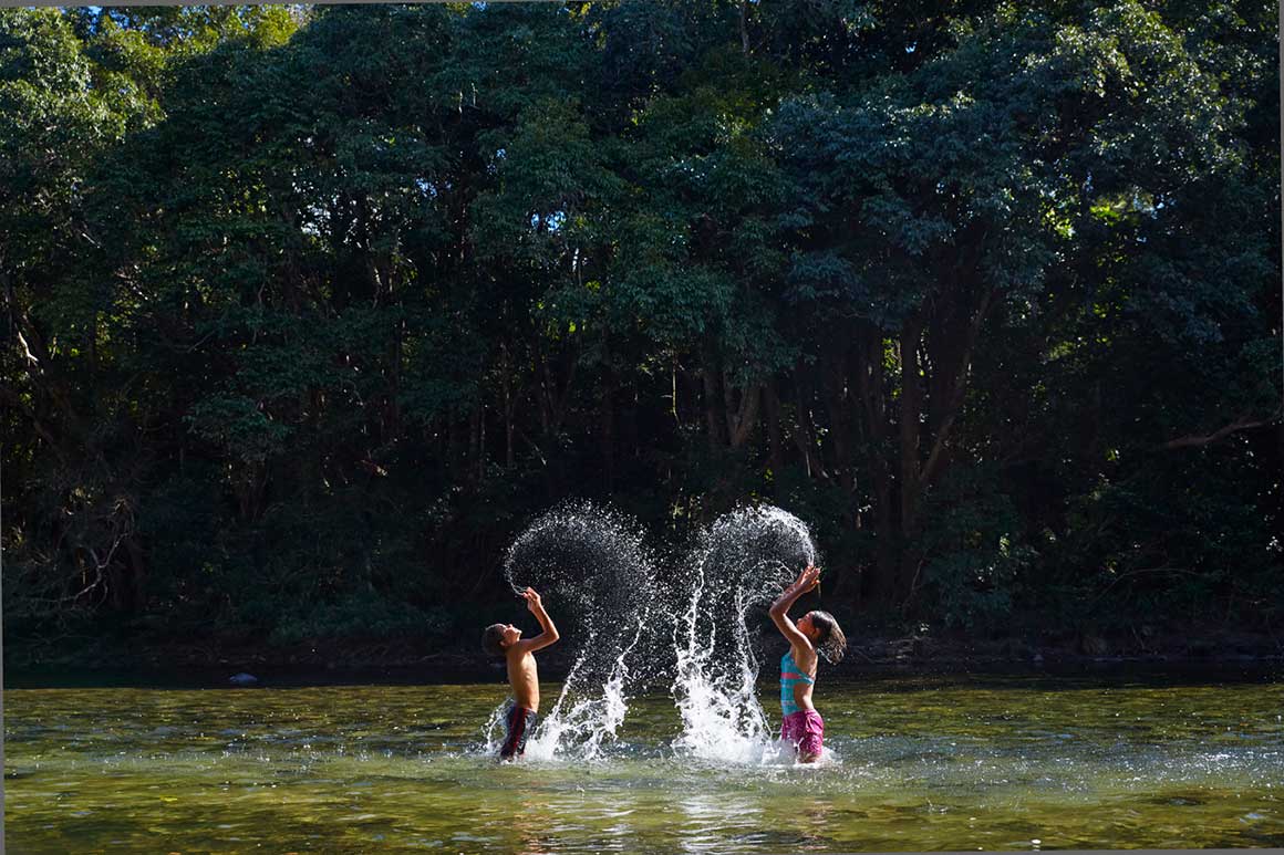 Two children splashing water into the air while standing in shallows of river with rainforest backdrop