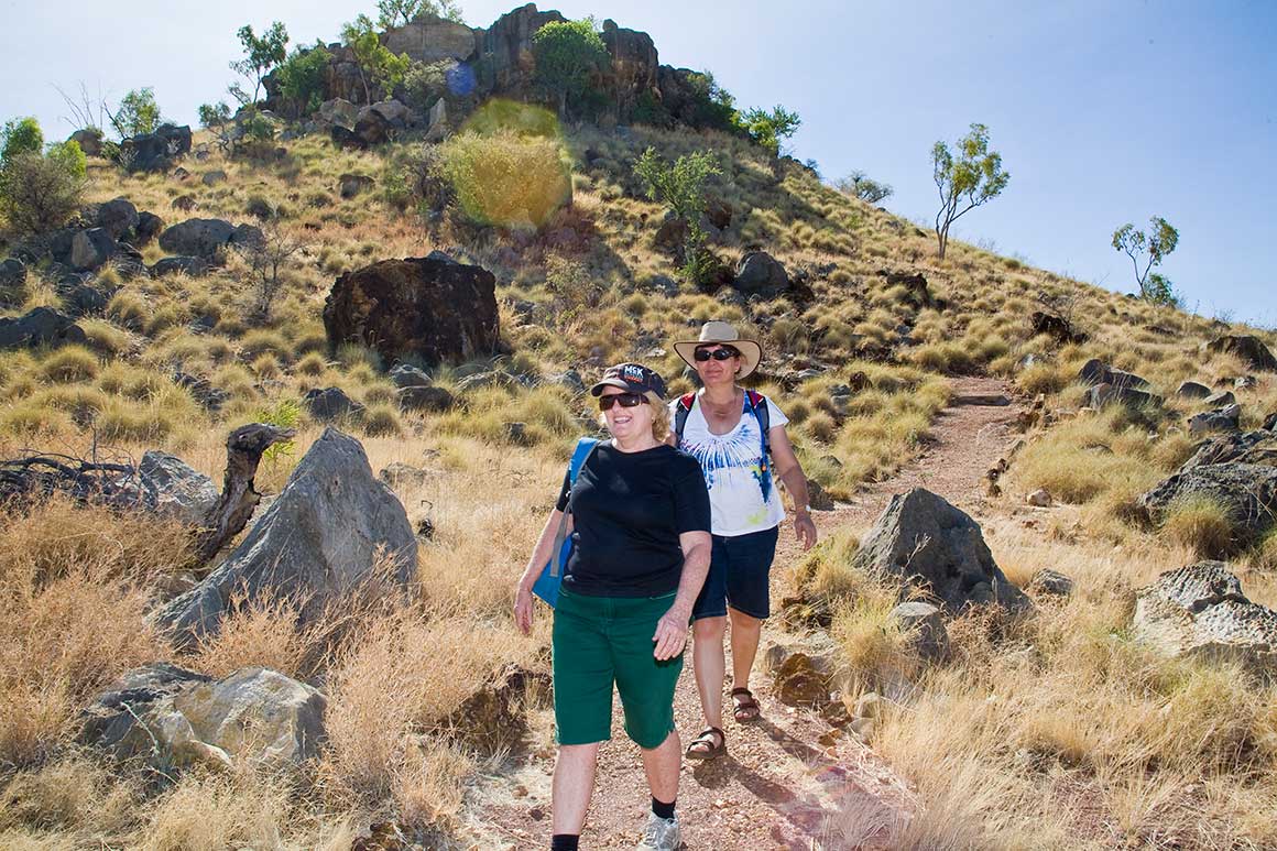 Two visitors walk along a rocky track winding down a gently sloping hill in a dry savanna landscape, with sparse trees and jagged rocks protruding above the yellow grass. 