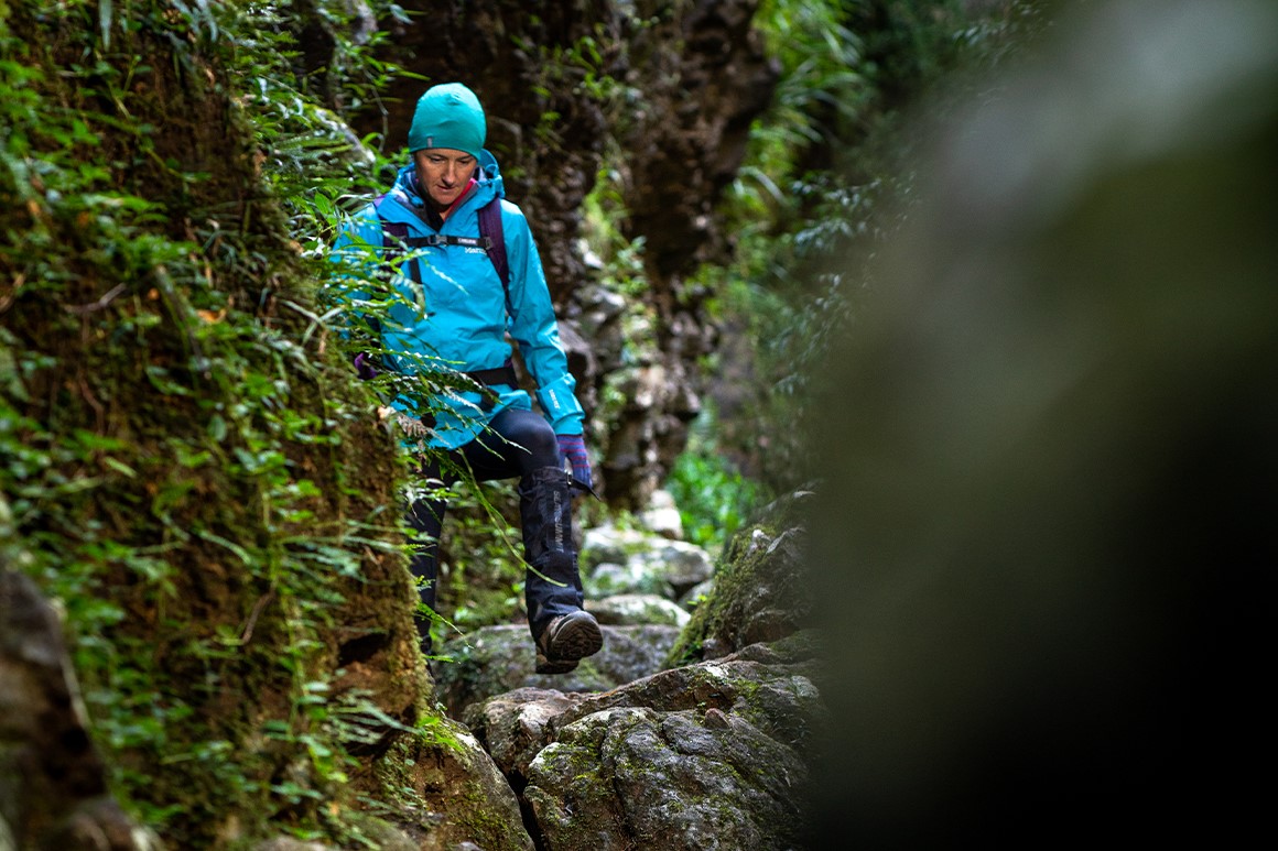 A female hiker in bright blue wet weather gear is walking across large rocks. 