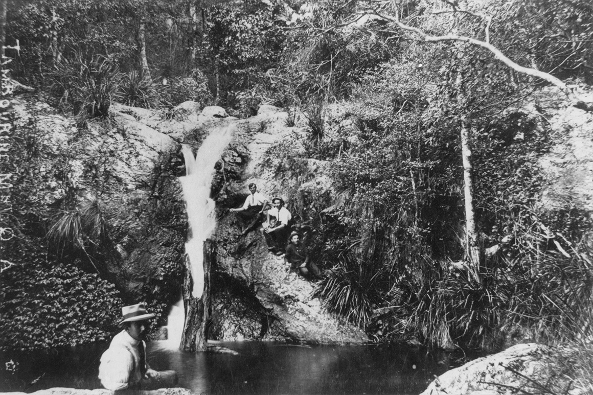 Old black and white image of people sitting on a rockface and around the rock pool underneath a waterfall surrounded by rainforest.