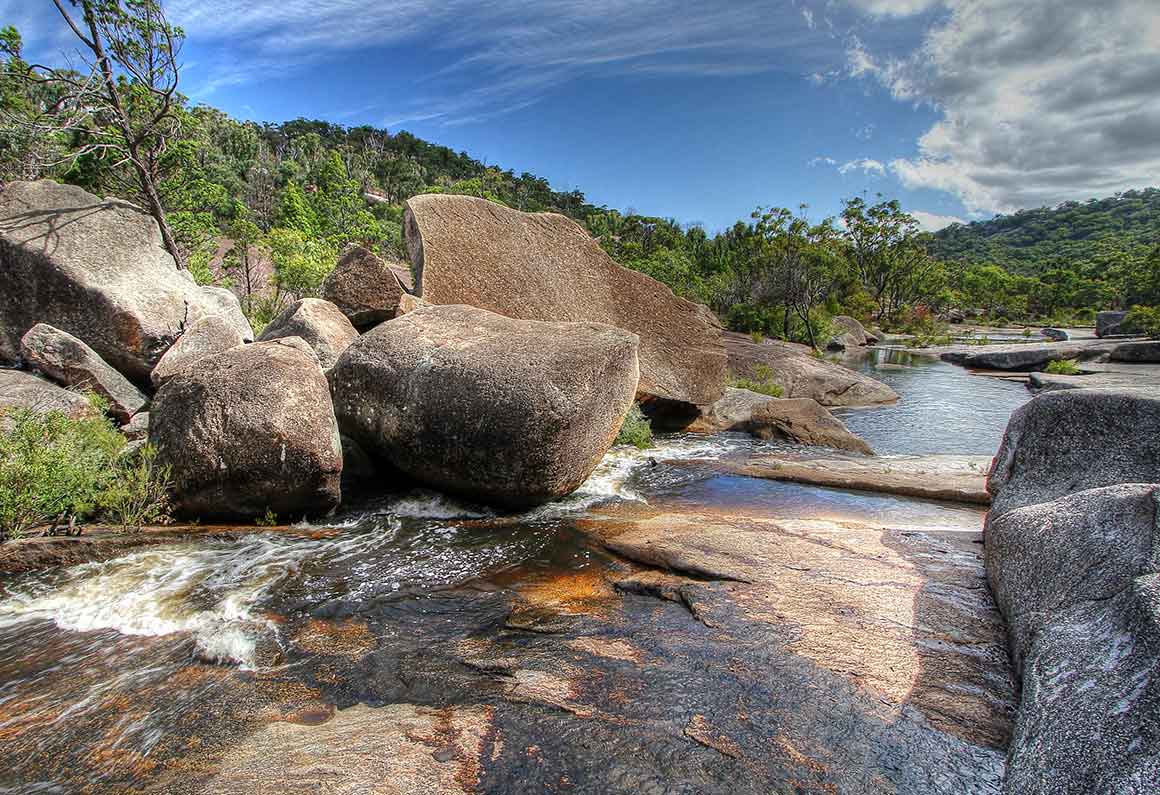 A shallow creek meanders across smooth granite rocks, with large boulders and forest on each side. 
