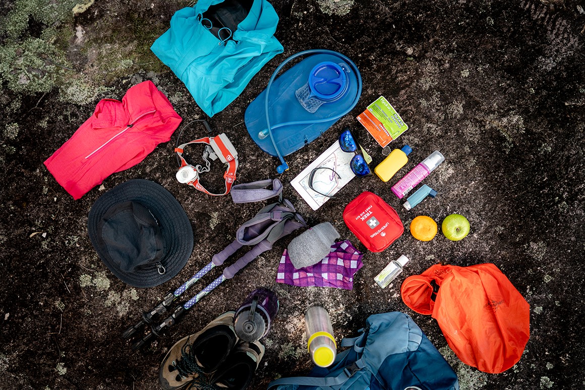 A selection of hiking equipment, including wet weather clothes, first-aid-kit, water bottle and headlight, is laid out neatly on a rock. 