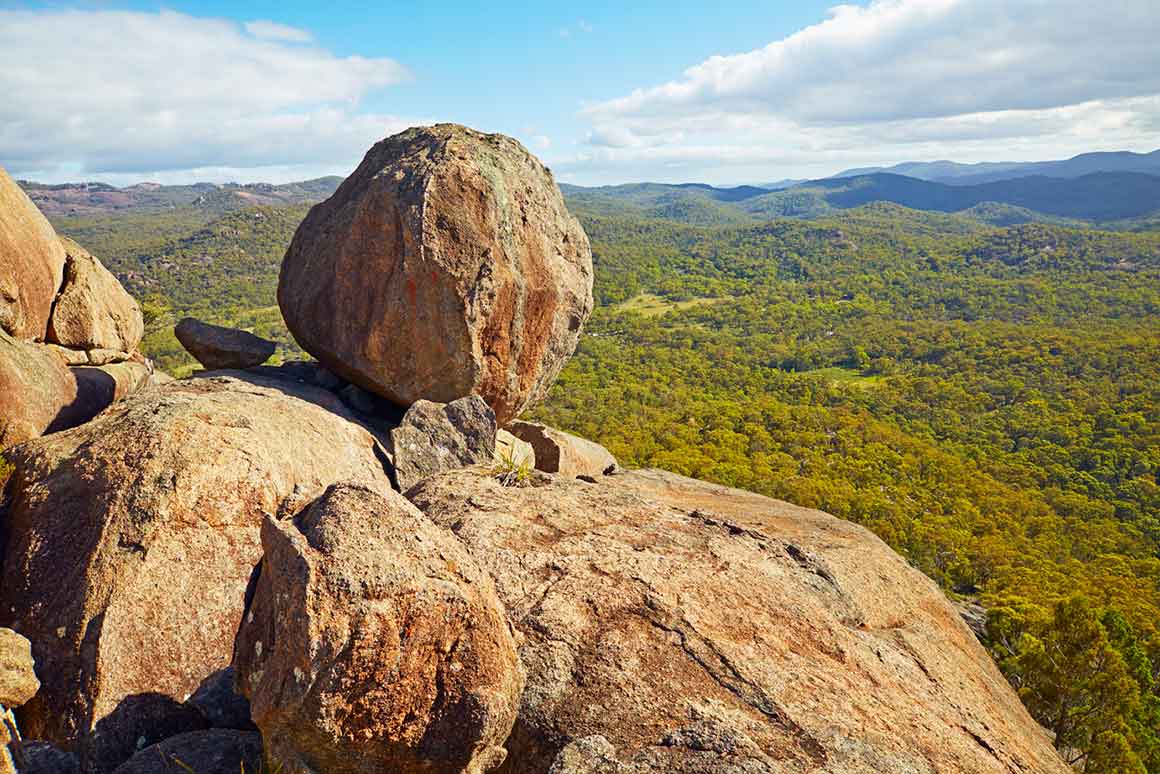 Huge granite boulders sit balanced on a massive granite outcrop, high above a forested valley with mountains in the distance.