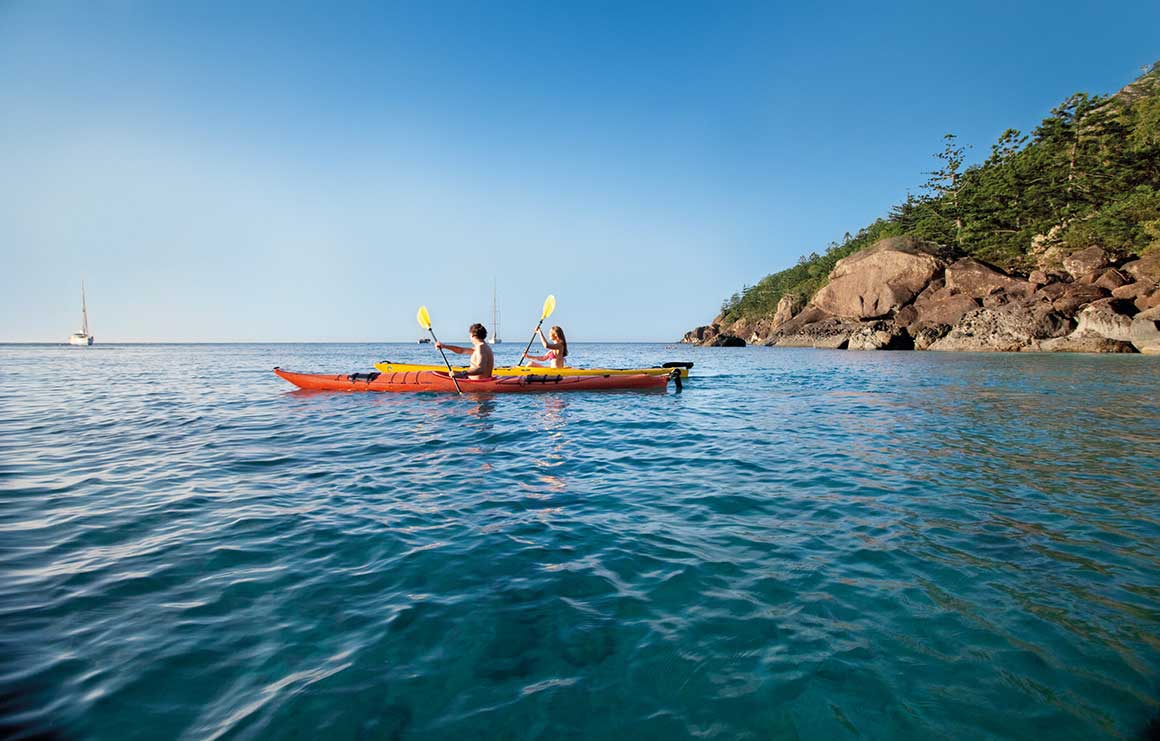 Two people paddling kayaks on a blue ocean with an island in the background.  