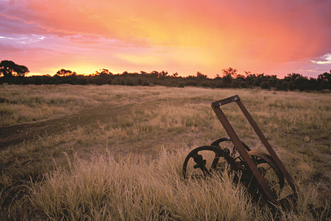 An old rusty farm implement sits in a paddock of dry grass against a backdrop of orange purple skies at sunset.
