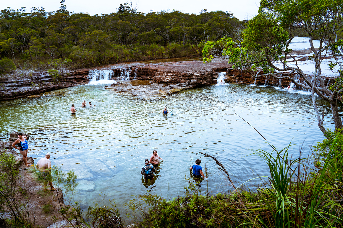Water cascades over a high rocky ledge into large rock pool where many people are swimming, surrounded by woodland. 