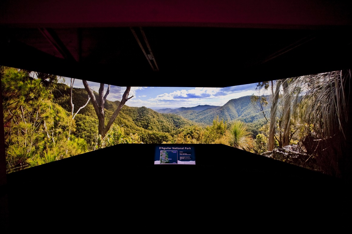 A wide screen angled in a semi-circle shows a panoramic image of forest in foreground and mountain ranges in background.