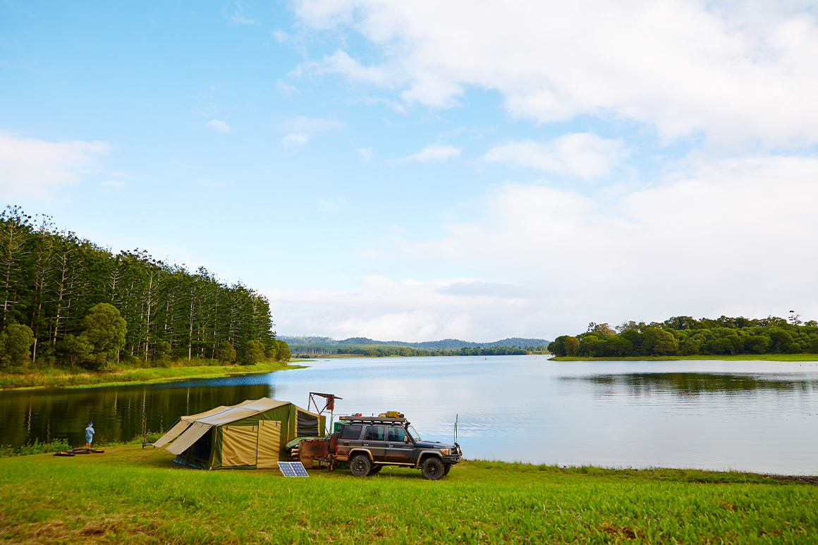 A four wheel drive vehicle is parked in front of a large tent, set on the grassy shore of a large forest-fringed lake, where the tranquil waters reflect the forest and blue skies above. 